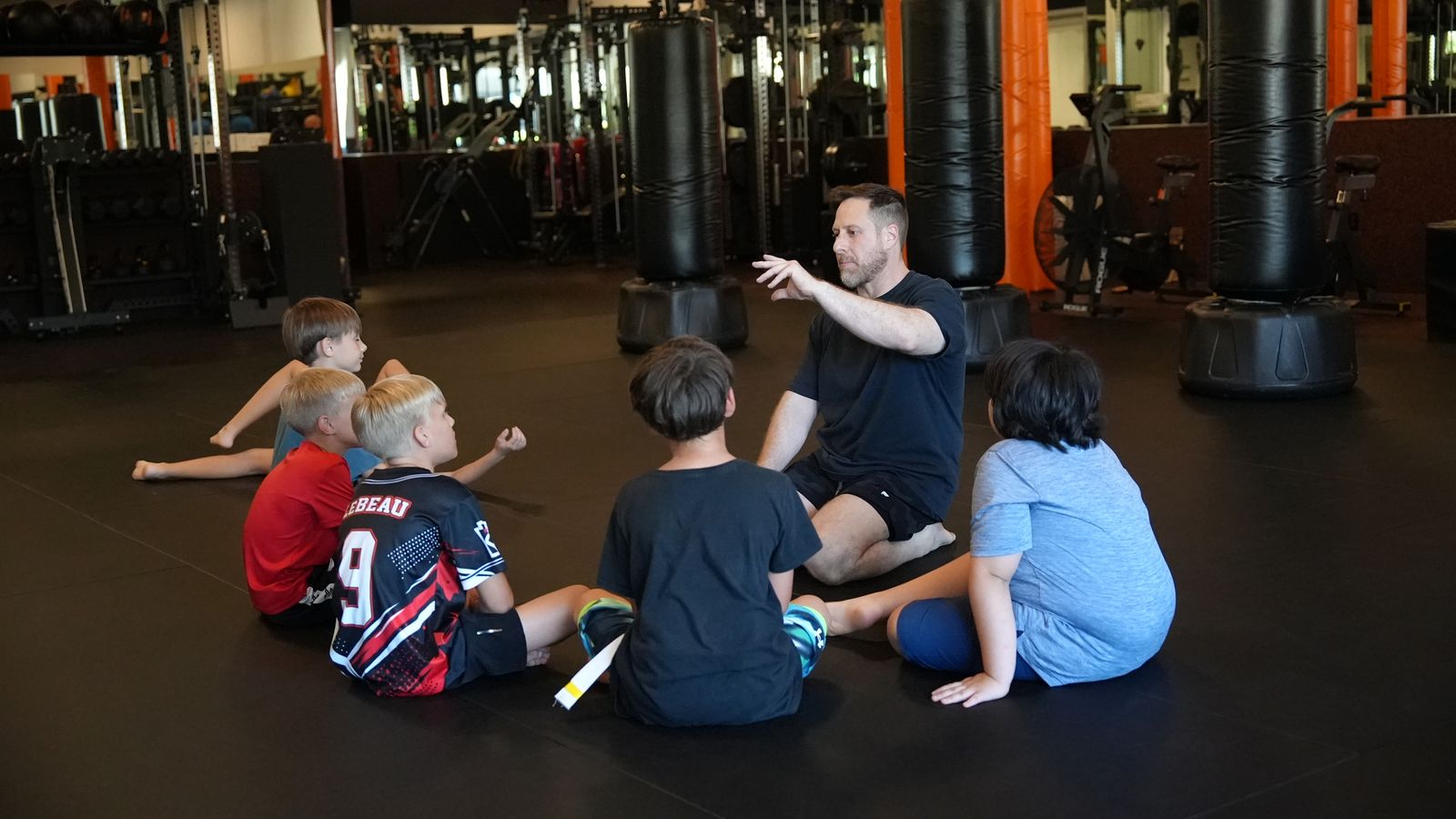 Coach Wil sits on the mat teaching a circle of young students at Forge Athletics in Naples