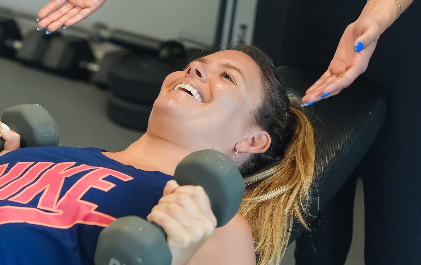 A member laughs mid-set as her coach spots a dumbbell press during personal training at Forge Athletics