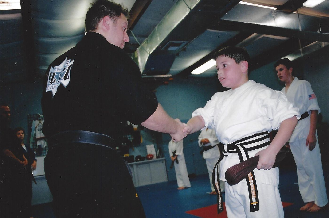 Wil Pino shakes hands with young student Michael Compagnone receiving his first-degree black belt at Elite Martial Arts